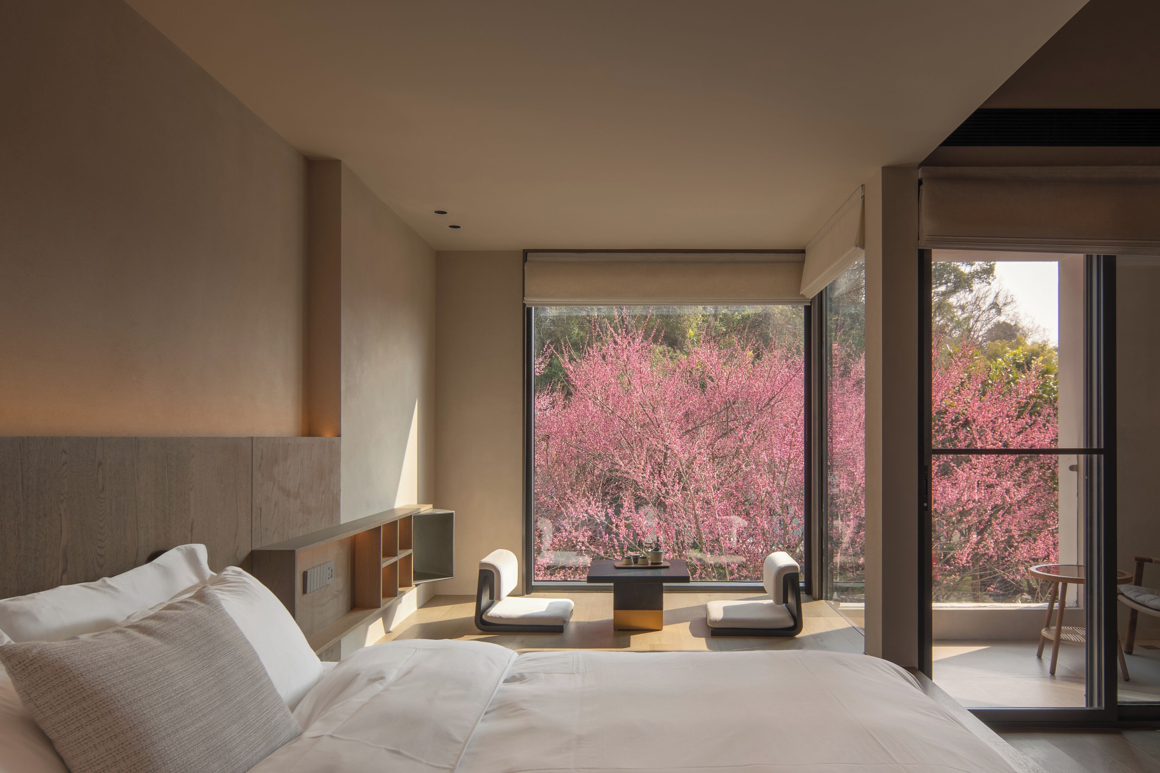 Un dormitorio moderno con una cama grande, una mesita pequeña, dos sillas y un balcón con vistas a los cerezos en flor.