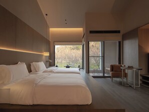 Ein modernes Hotelzimmer mit zwei Betten, einem Schreibtisch, einem Sessel und großen Fenstern mit Blick auf Bäume.