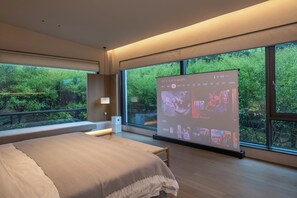 Ein modernes Schlafzimmer mit einem großen Bett, einer Projektionsleinwand, die Bilder zeigt, und Blick ins Grüne.