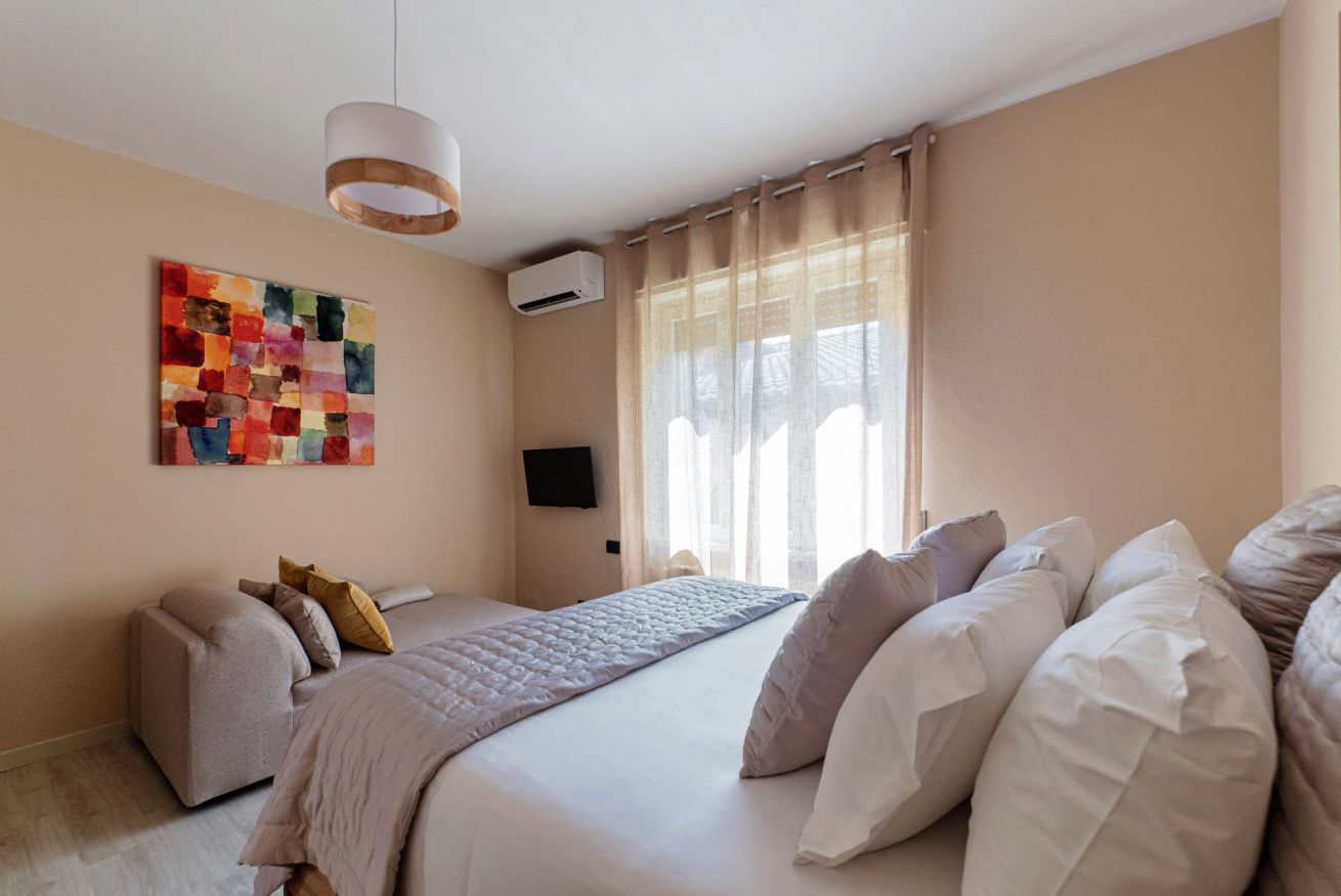 Quarto com cama, TV fixada na parede, luz no teto e janela com cortinas.