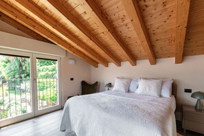 Ein Schlafzimmer mit Holzbalkendecke, einem Bett mit weißer Strukturdecke, einem Nachttisch mit Lampe und einer Glastür zum Außenbereich.