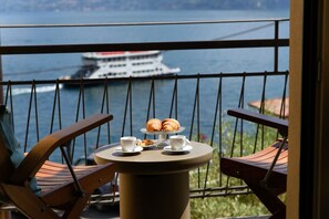 Een balkon met twee stoelen, een kleine ronde tafel met croissants en kopjes, en uitzicht op een cruiseschip op het water.