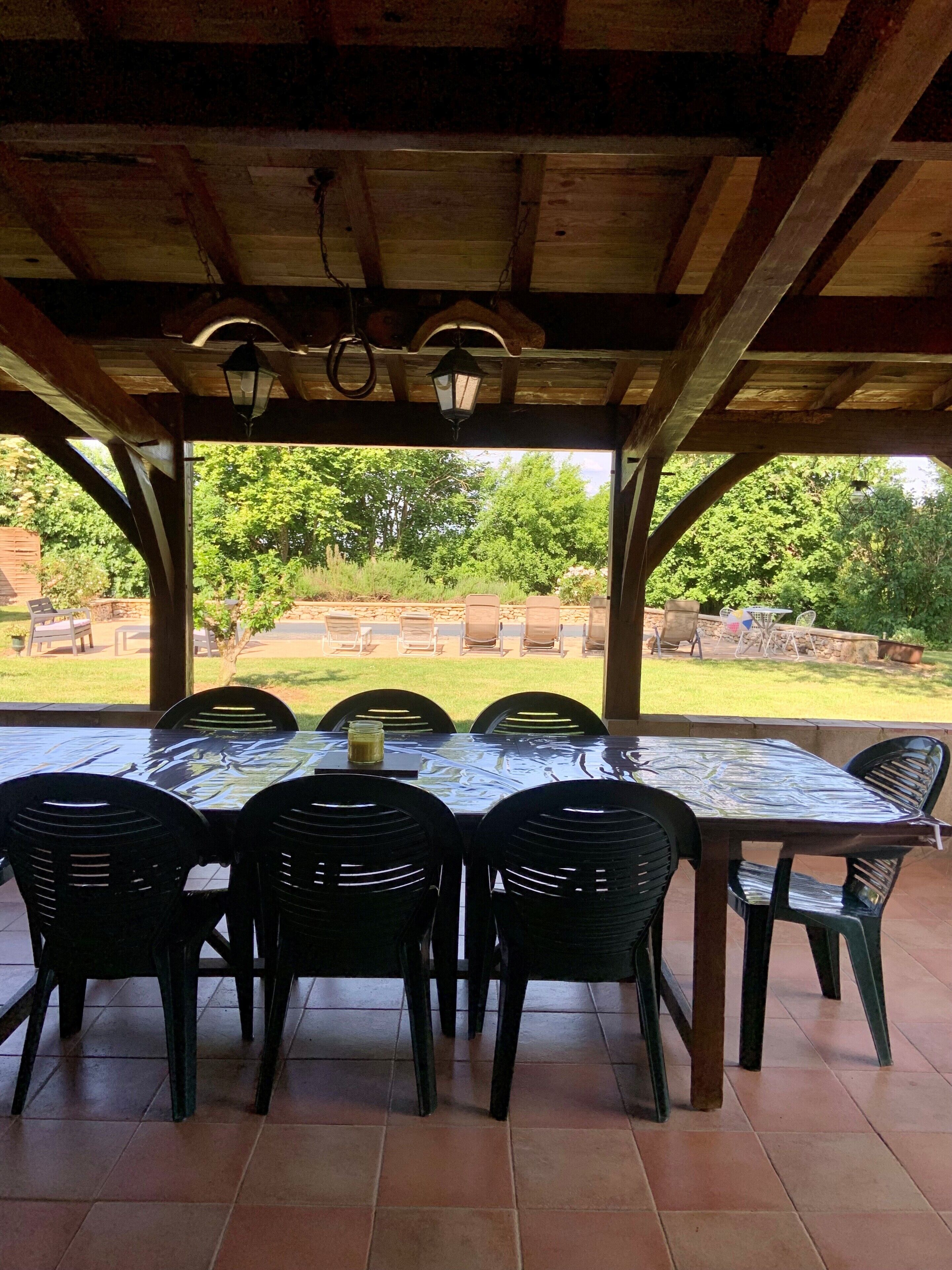 En udendørs overdækket terrasse med et bord, stole, en lygte og udsigt til en have.