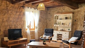 A rustic living room with stone walls, a wooden ceiling, a television on a wooden stand, a sofa, two armchairs, a coffee table, and a bookshelf. - Country house in Périgord with swimming pool (Beaumontois en Périgord)