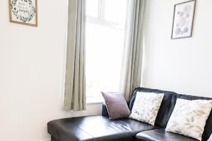 A room with a black leather sofa, two pillows, a window with curtains, and two framed pictures on the wall. - House Sleeps 7 With Wifi (Gateshead)