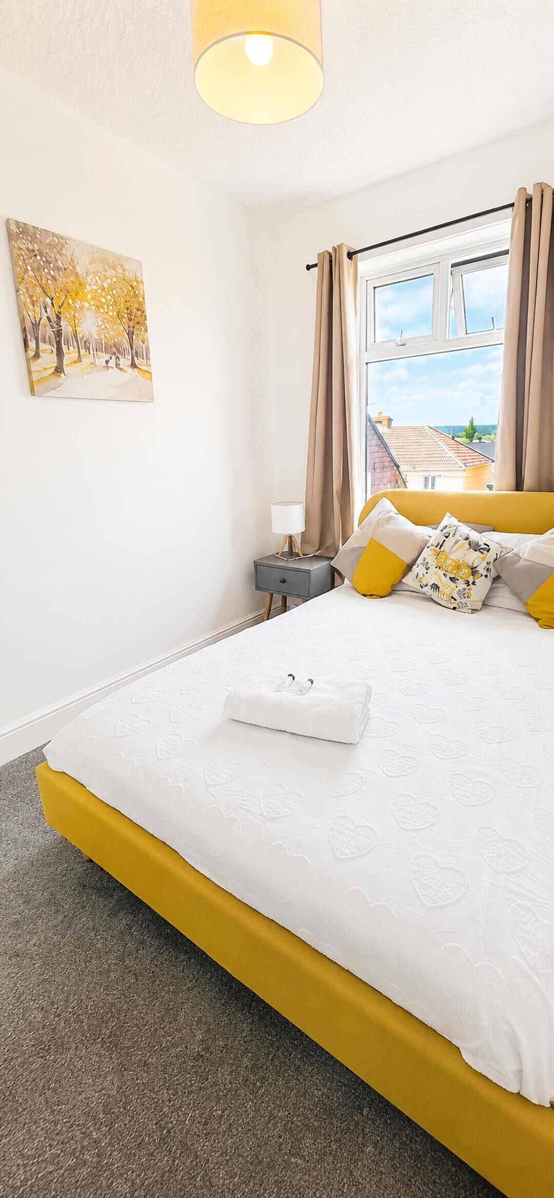 Een slaapkamer met een bed, een nachtkastje, een lamp, een schilderij en een raam met gordijnen.