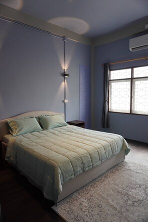 A bedroom with a bed, a nightstand, a wall-mounted lamp, a window with curtains, and an air conditioning unit. - Large Room Near Grand Palace (Bangkok)