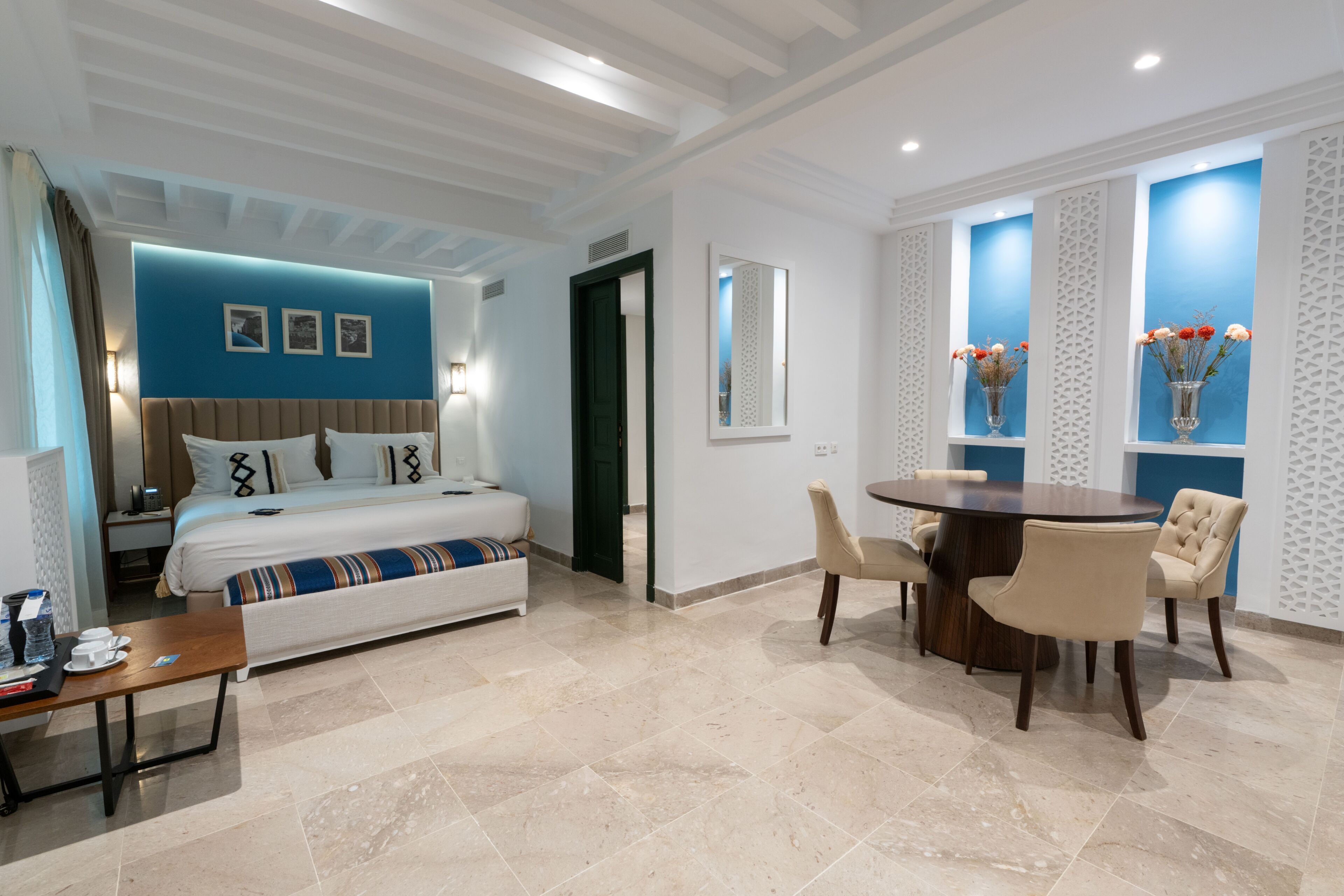 Una habitación de hotel con cama, mesita de noche, lámpara y un área de comedor con sillas y una mesa.