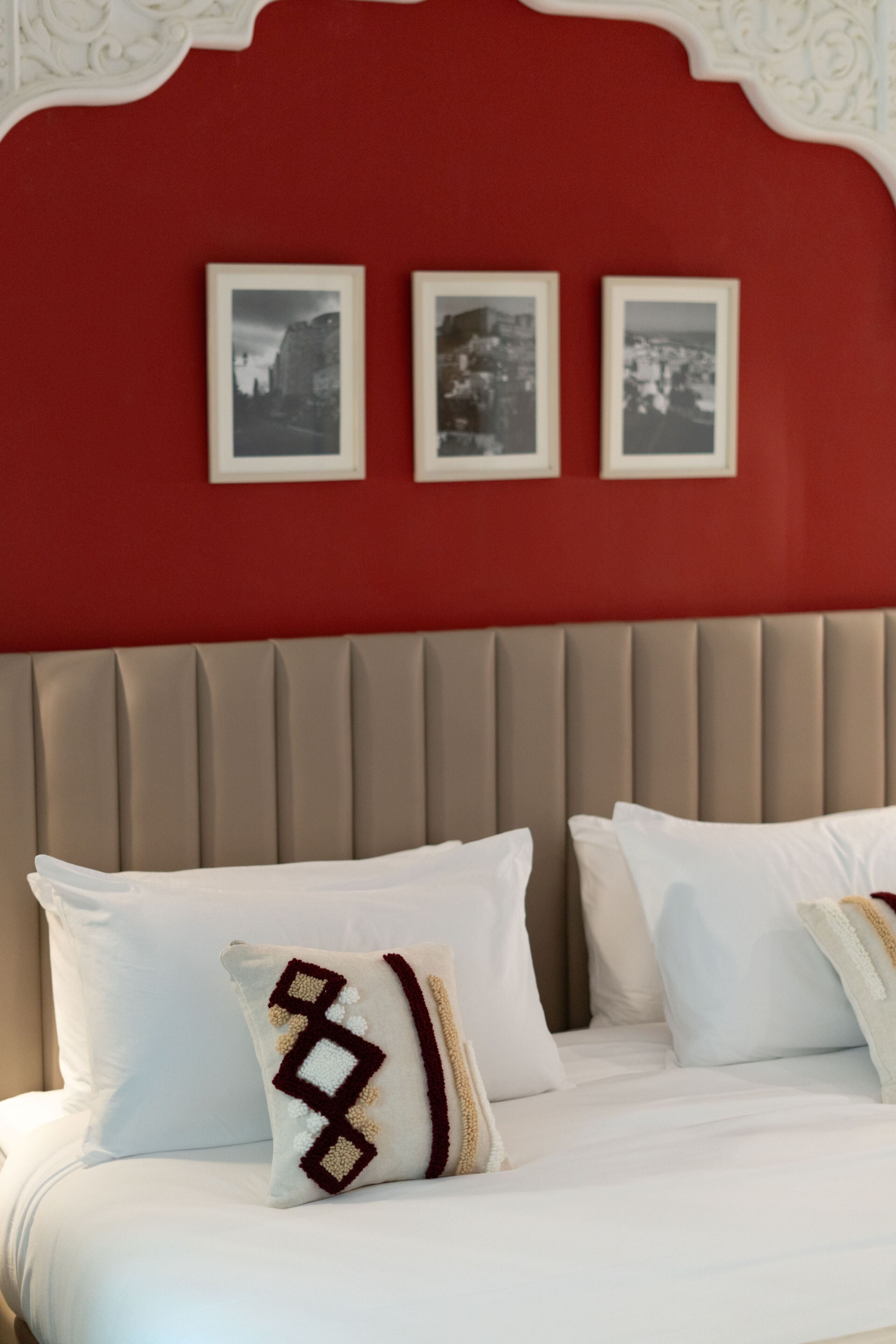 Una cama con ropa blanca y una almohada estampada, una pared roja con cuadros enmarcados y un cabecero color beige.