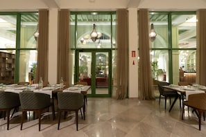 Un comedor con mesas dispuestas para cuatro, una araña y vistas a través de un ventanal hacia otro salón comedor.