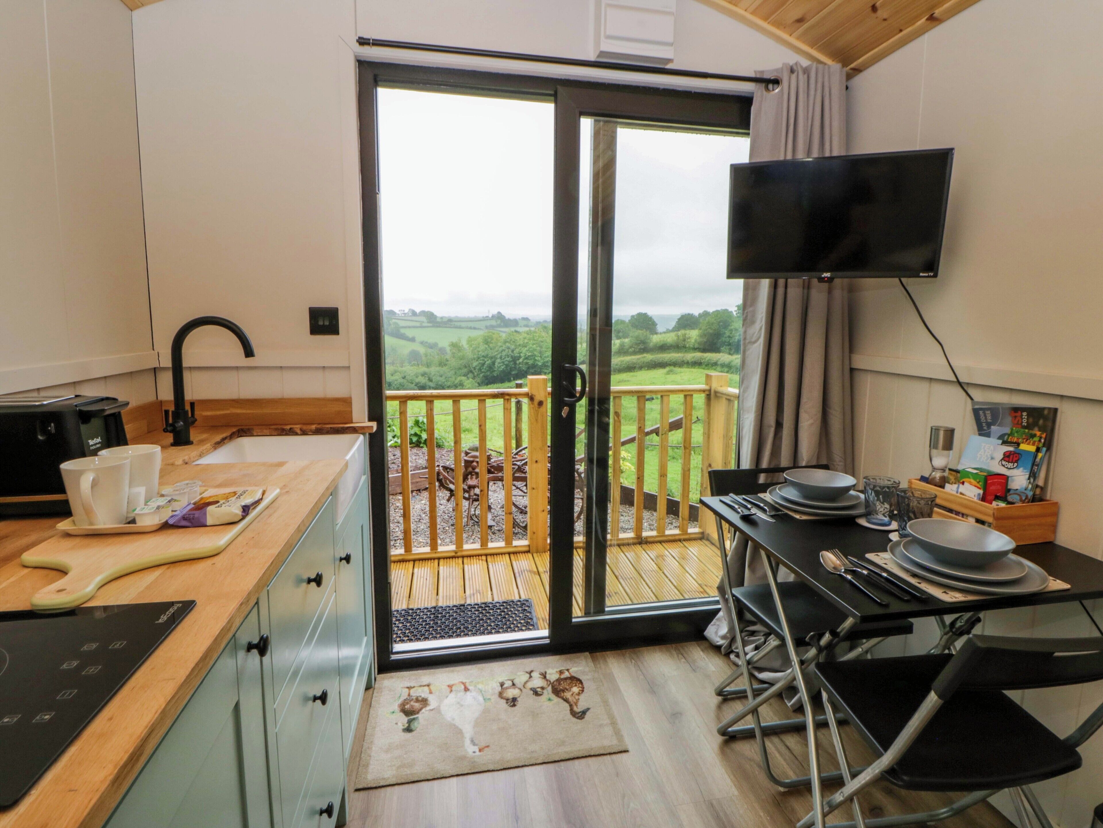 A kitchenette with a toaster, a coffee maker, and a cutting board. There is a dining area with a table, chairs, and a TV mounted on the wall. A sliding glass door leads to a balcony with a view of the countryside.