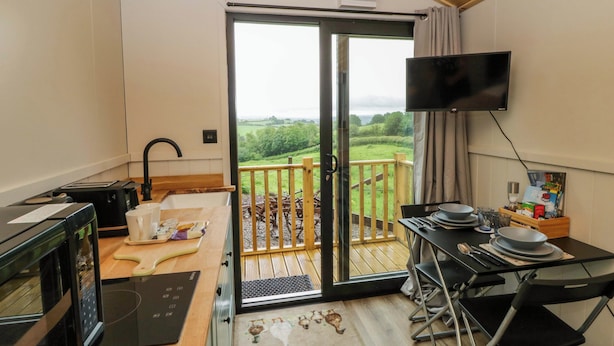 A kitchen with a microwave, toaster, and sink, a dining area with a table and chairs, a TV mounted on the wall, and a balcony with a view of the countryside. - The Hut (Llandysul)