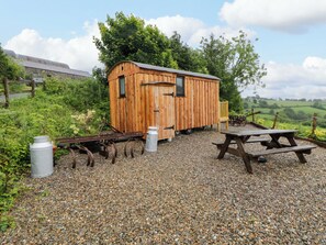 Ein hölzernes Gartenhaus mit Tür und Fenstern, eine Schotterfläche mit Picknicktisch und eine Metallkonstruktion.