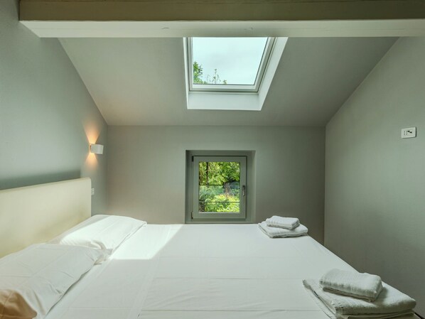 A bedroom with a skylight, a bed with white linens, a small window, and a wall-mounted light.