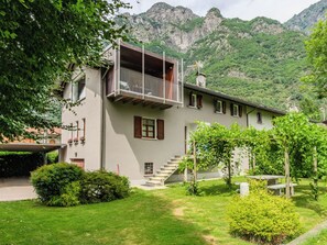 Ein zweistöckiges Gebäude mit einem Balkon, einer Treppe, die zum Eingang führt, und einem Garten mit Sträuchern und einer Bank.