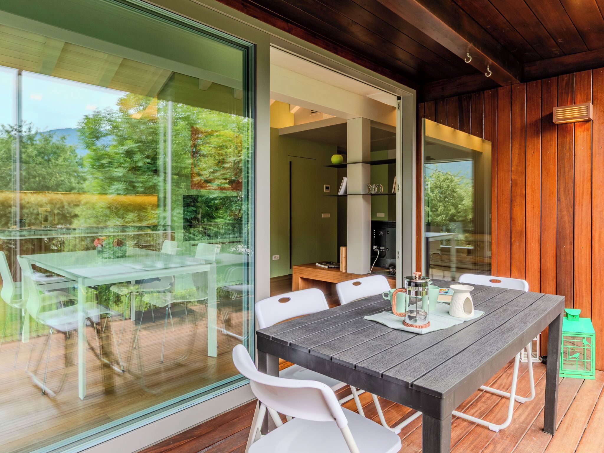 Un espace de restauration en plein air avec une table en bois et des chaises blanches, une cloison en verre et une vue sur la verdure.
