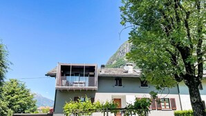 Uma casa de dois andares com varanda, jardim e montanha ao fundo.