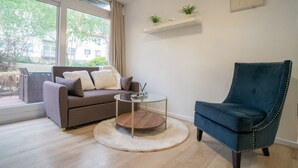 Ein modernes Wohnzimmer mit einer Couch, einem runden Couchtisch, einem blauen Sessel, einem weißen Teppich und einem Fenster mit Blick auf Grünflächen.