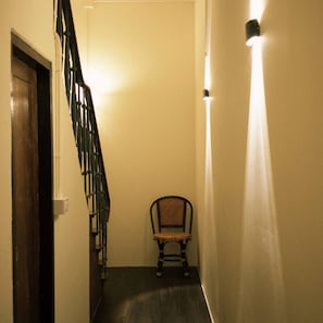 A narrow hallway with a wooden staircase on the left, a chair in the center, and a wall-mounted light on the right. - Family Room Near Chinatown (Bangkok)
