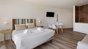 A modern hotel room with a bed, a desk, chairs, a TV, and a kitchen area.