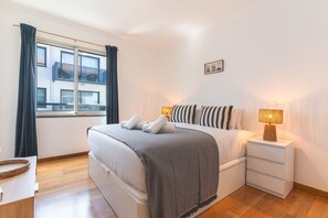 Ein ordentlich eingerichtetes Schlafzimmer mit Bett, Nachttischen, einem Fenster mit Vorhängen und Blick auf Gebäude.