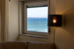Ein Fenster mit Blick auf das Meer und eine Stadtlandschaft, eine Wandleuchte und ein Teil eines Raumes mit einem braunen Sessel.