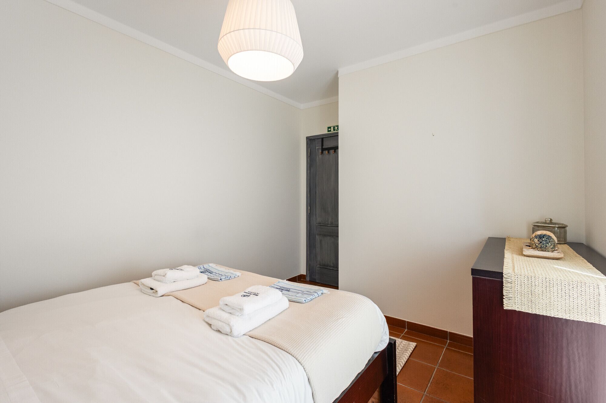 A hotel room with a bed, a desk with a lamp, and a door.