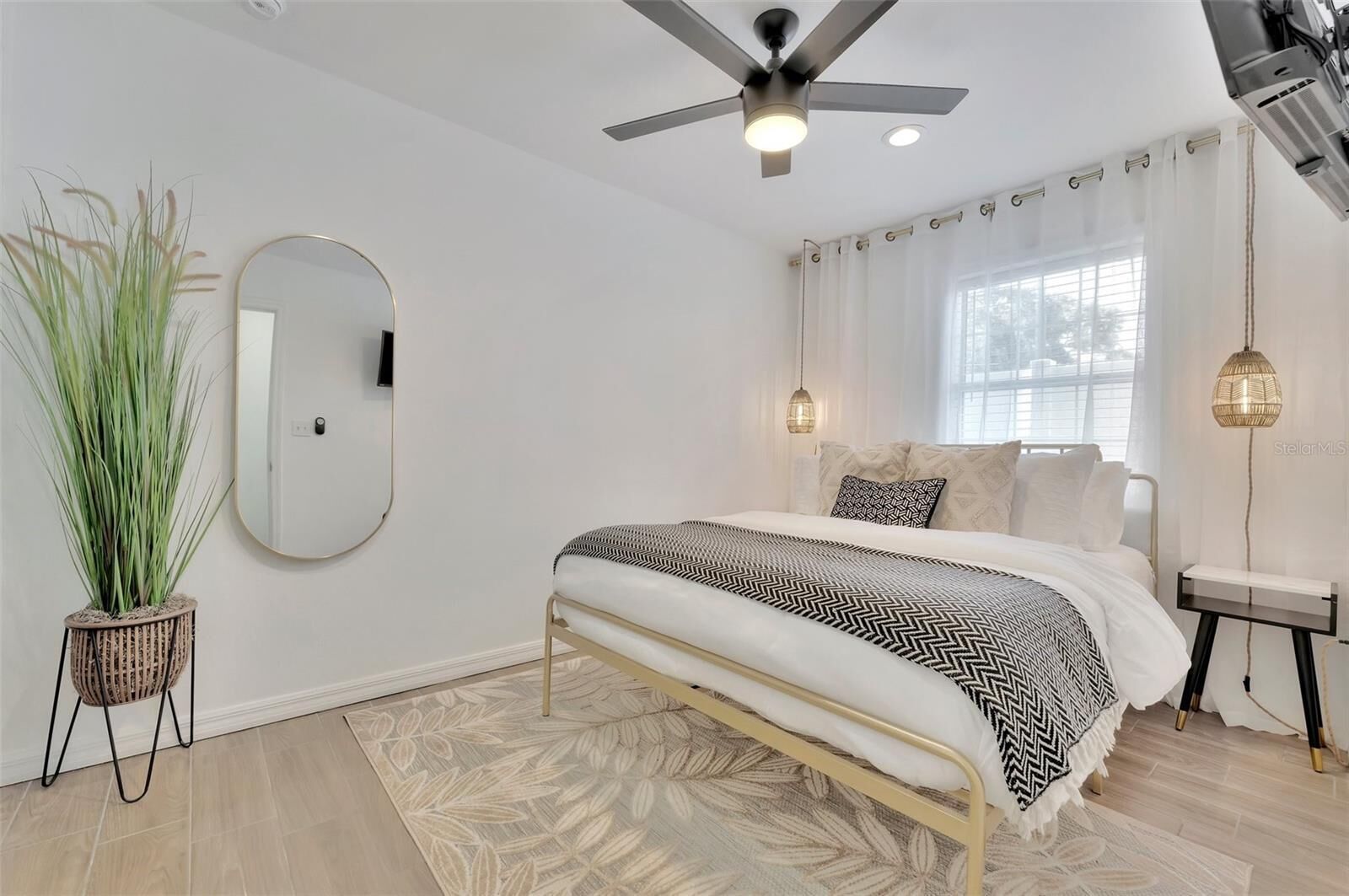 Un dormitorio con cama, ventilador de techo, espejo grande, planta en maceta y ventana con cortinas.