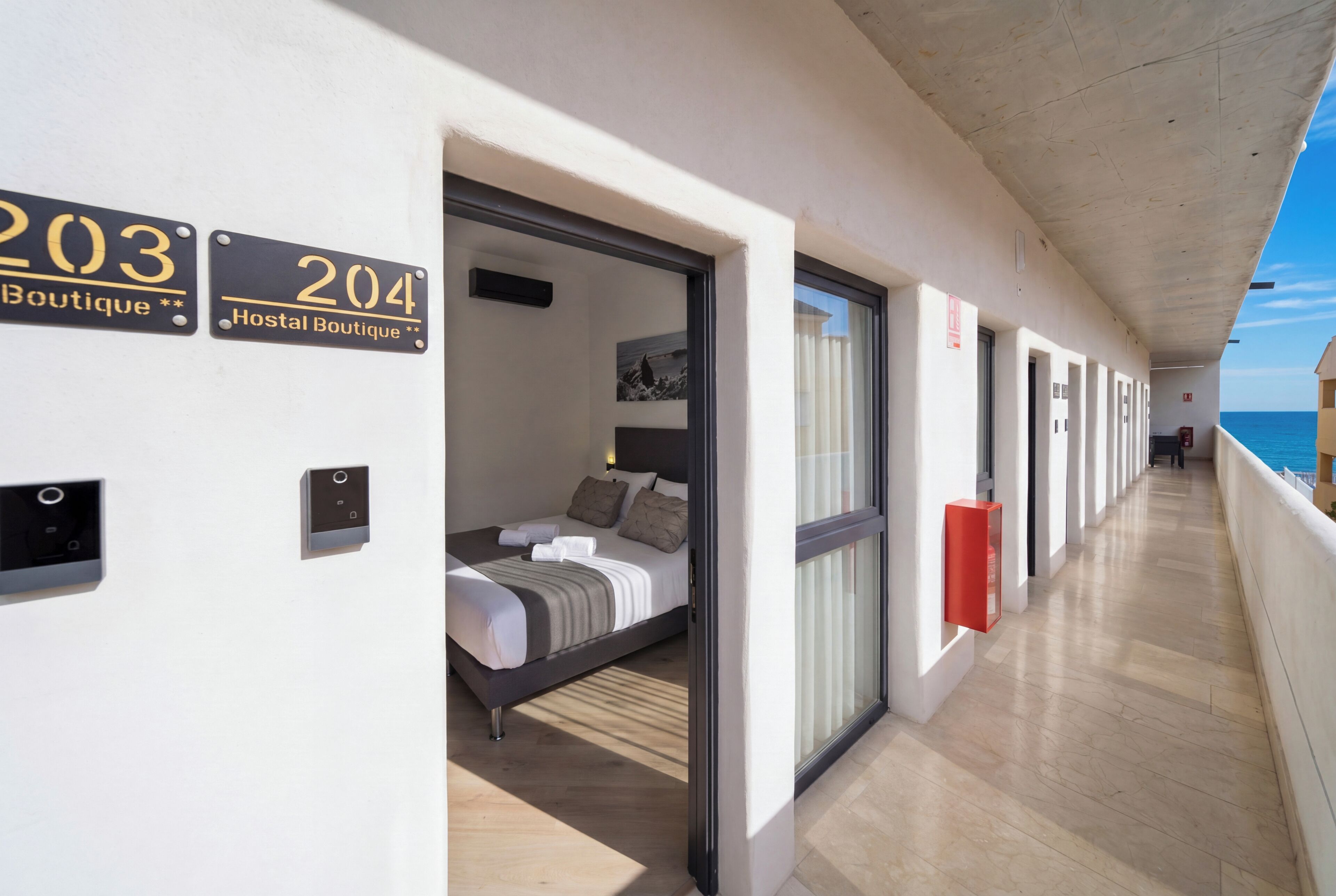Ein Hotelgang mit nummerierten Zimmern, einem Bett, einer Schiebetür, einem Feuerlöscher und Meerblick.