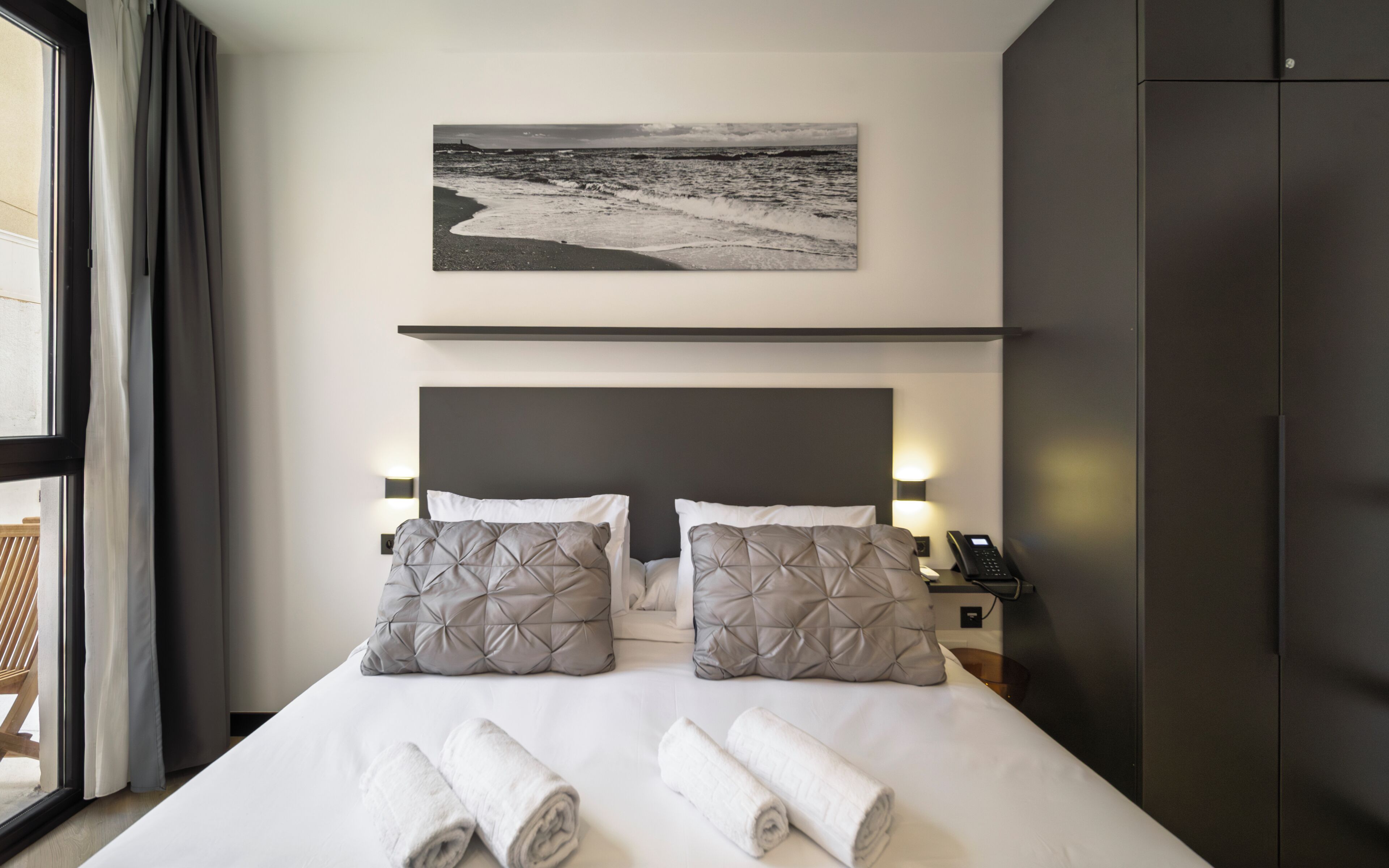 Una habitación de hotel con una cama, dos almohadas, una mesita de noche con un teléfono, un cuadro en la pared y una ventana con cortinas.