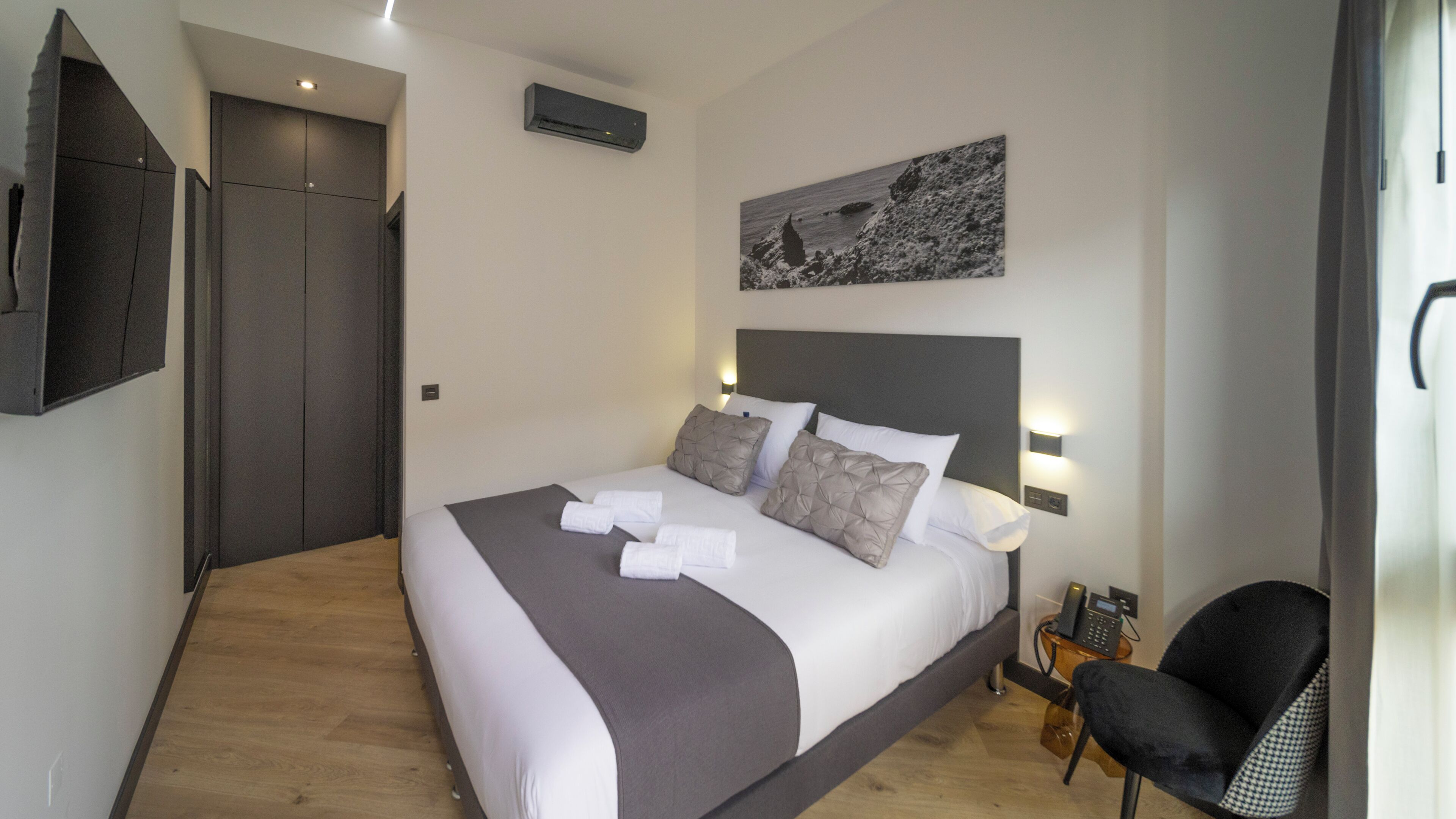 A hotel room with a bed, a chair, a bedside table with a phone, a painting on the wall, and a door.
