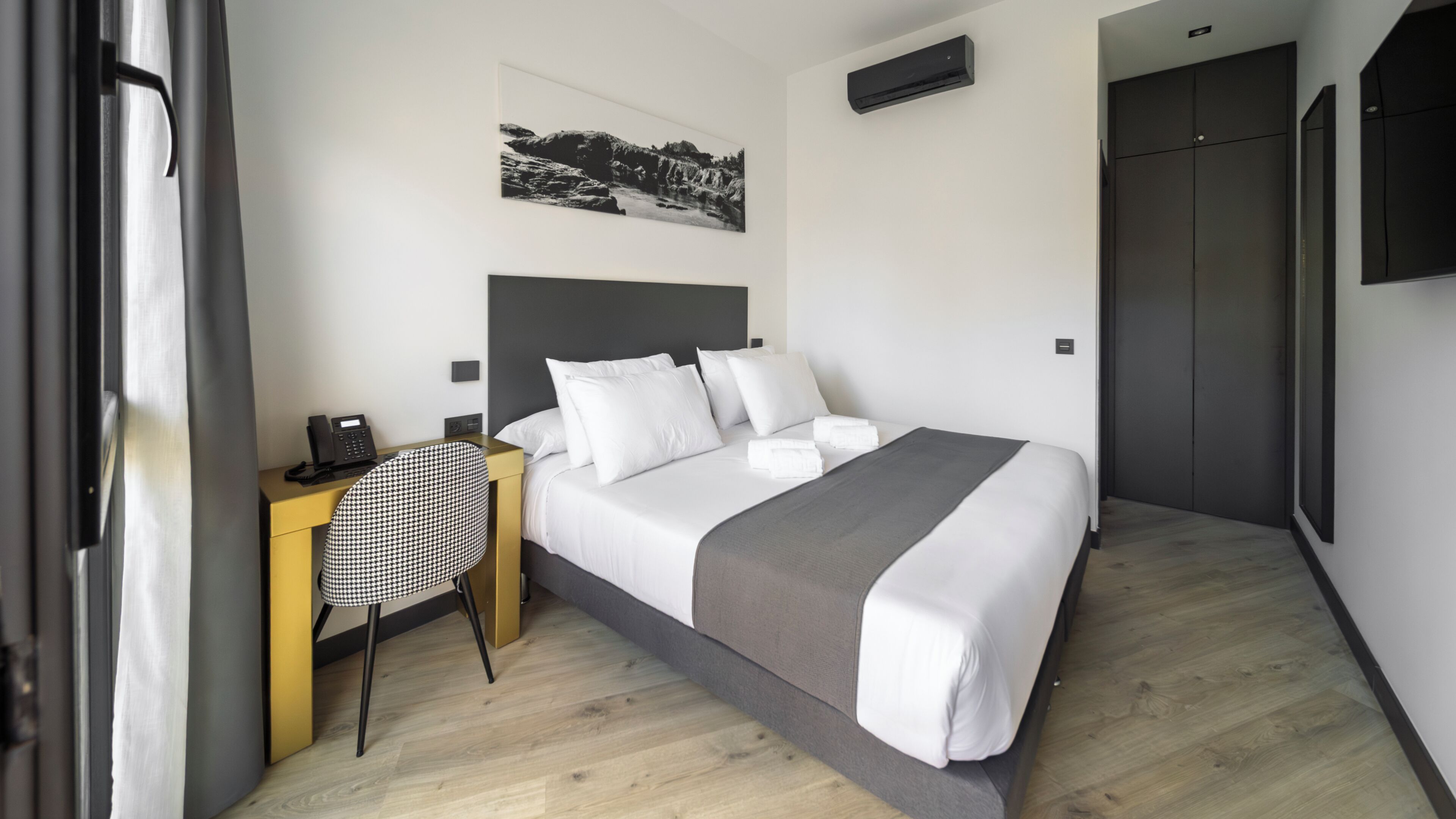 A hotel room with a bed, a desk with a telephone, a chair, a painting on the wall, and a wardrobe.