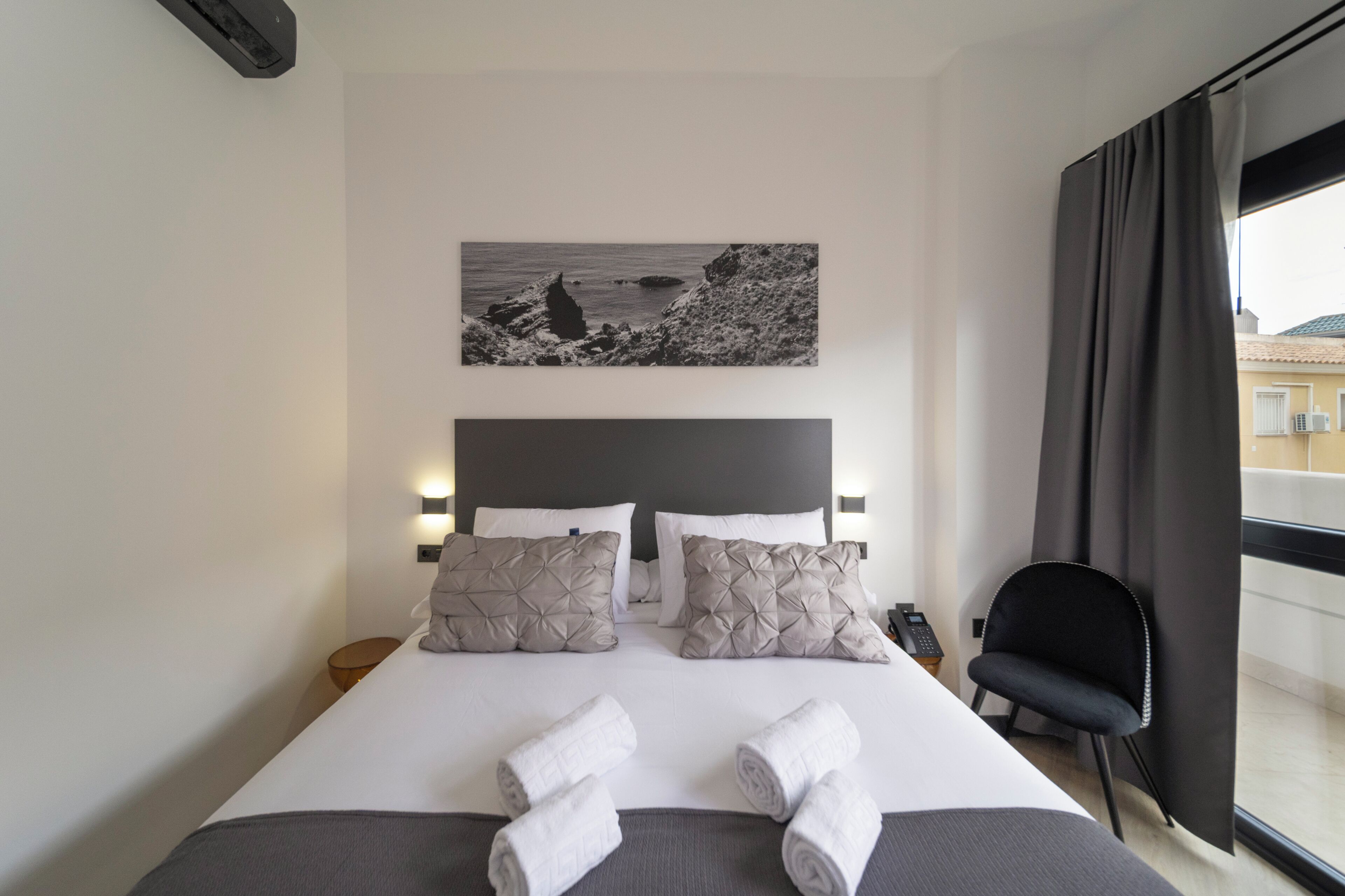 A hotel room with a bed, pillows, a bedside table, a chair, a window, and a wall-mounted artwork.