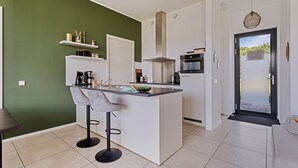 Een moderne keuken met een centraal eiland, barkrukken, een inbouwoven en een deur met een zwart frame die leidt naar een balkon.