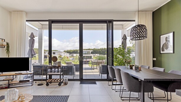 Een moderne woonkamer met een grote televisie, een eettafel en stoelen, een schuifdeur naar een terras buiten, en een hanglamp.