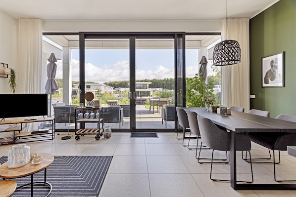 Una sala moderna con televisor grande, mesa de comedor y sillas, puerta corrediza de vidrio que da a un patio exterior y una lámpara colgante.