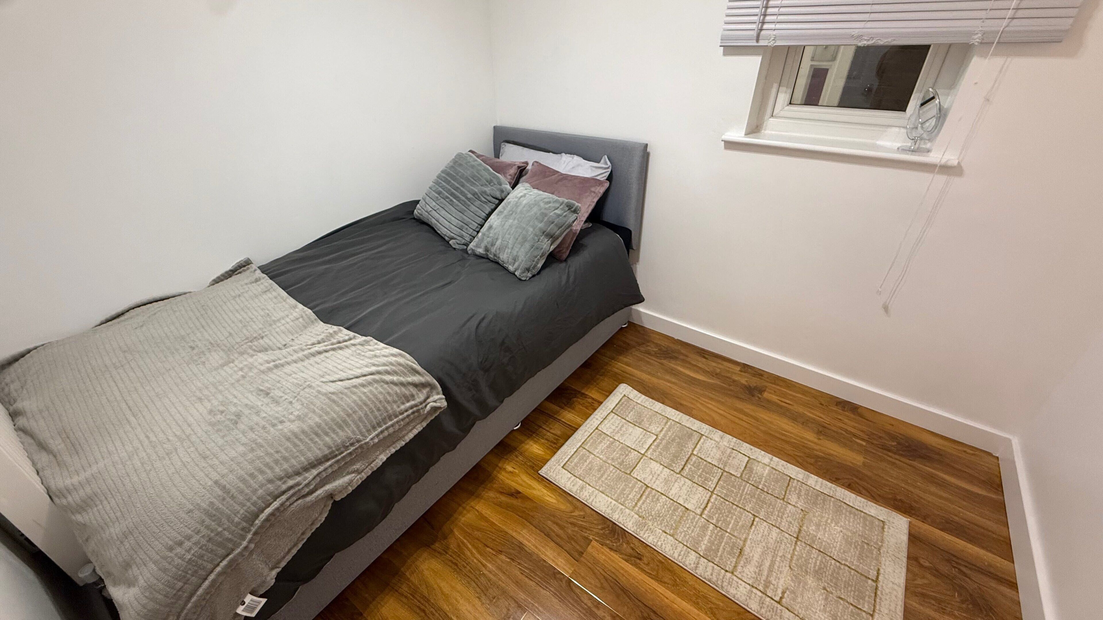 A small, single-bed room with a grey headboard, a window with blinds, a wooden floor, and a small rug.