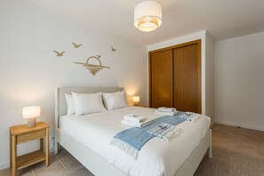 Ein ordentlich bezogenes Bett mit weißen Leinen, ein Nachttisch aus Holz mit einer Lampe, eine Wandleuchte und ein Holzschrank.