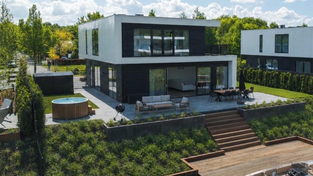 Una casa moderna de dos pisos con un patio, mobiliario exterior y un muelle con un bote.