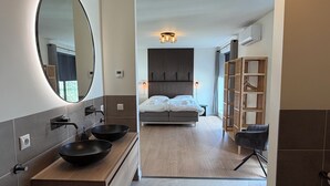 Un baño moderno con un espejo grande, dos cuencos negros, un mueble de tocador de madera y una cama con vistas al exterior.