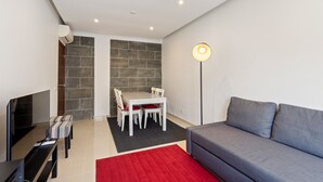 Uma sala de estar moderna com um sofá cinzento, um tapete vermelho, uma área de jantar com mesa e cadeiras, e uma televisão.