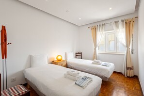 Un dormitorio con dos camas, una mesita de noche con lámpara, una silla, un banco y una ventana con cortinas.