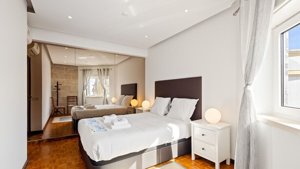 Quarto de hotel moderno com cama grande, mesas de cabeceira, espelho e janela com cortinas.