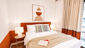 Et hotelværelse med en seng, et natbord med lampe, et billede i ramme på væggen og et vindue med gardiner.