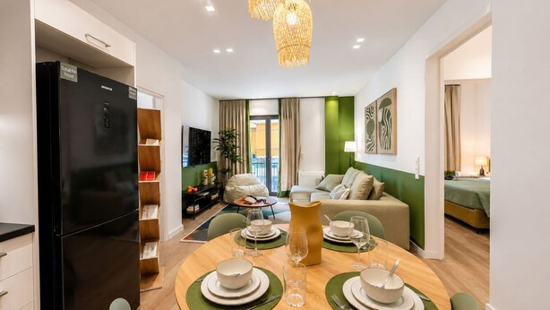 Une cuisine moderne avec un réfrigérateur noir, une table à manger pour quatre personnes et un coin salon avec un canapé et un téléviseur.