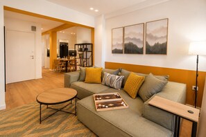 Ein modernes Wohnzimmer mit einer Couch, einem Couchtisch und einem Schachbrett. Im Hintergrund ist ein Essbereich mit Tisch und Stühlen zu sehen.