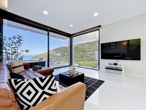 A modern living room with a large TV, a sofa with patterned cushions, a coffee table, and a view of a hillside. - Pearl of Madeira by Atlantic Holiday (Calheta)