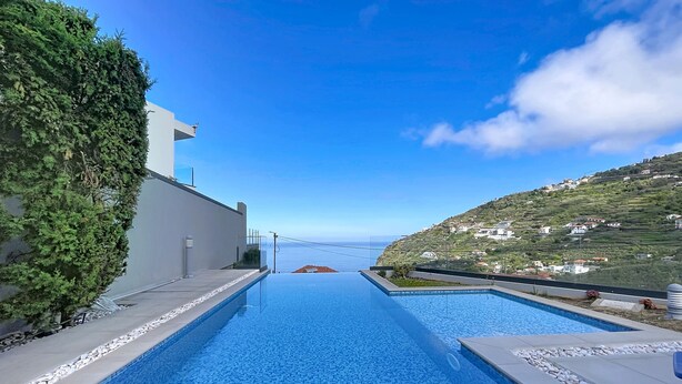 A swimming pool with clear blue water, surrounded by a white building and a hillside with houses. - Pearl of Madeira by Atlantic Holiday (Calheta)