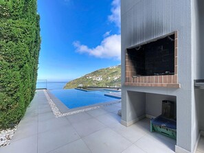 A modern outdoor pool area with a clear view of the sea and a hillside in the background. - Pearl of Madeira by Atlantic Holiday (Calheta)