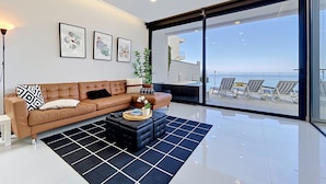 A modern living room with a brown leather sofa, a black ottoman, and a black and white rug. There are three framed artworks on the wall, a potted plant, and a view of the ocean through large glass doors. - Pearl of Madeira by Atlantic Holiday (Calheta)